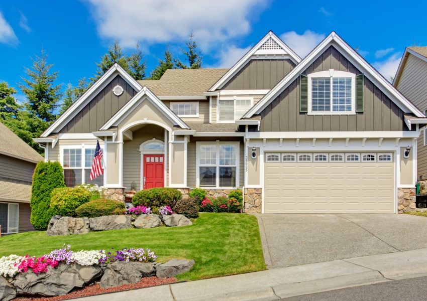 Grey house exterior with entrance porch and red door. Beautiful front yard landscape with vivid flower and stones